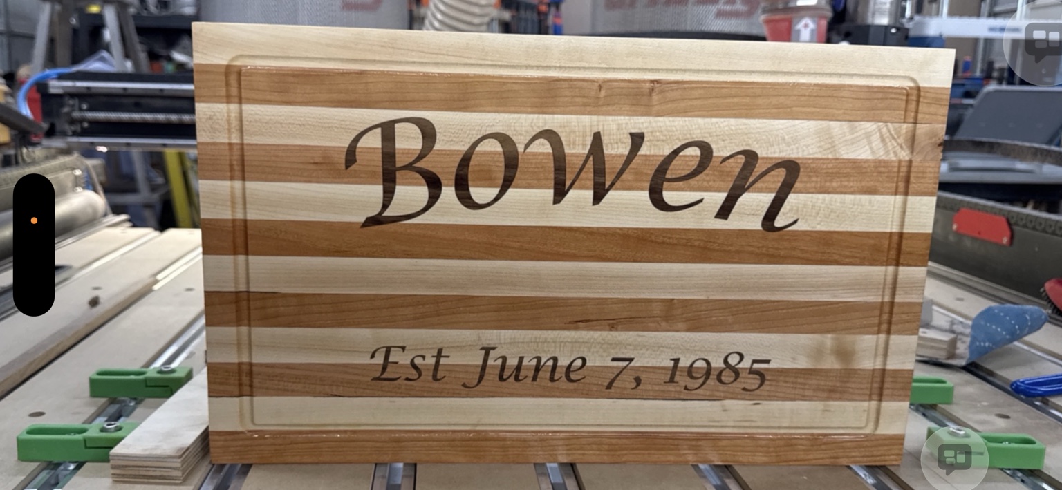 Close view of the actual Bowen engraved cutting board on the CNC table, with date and juice groove visible
